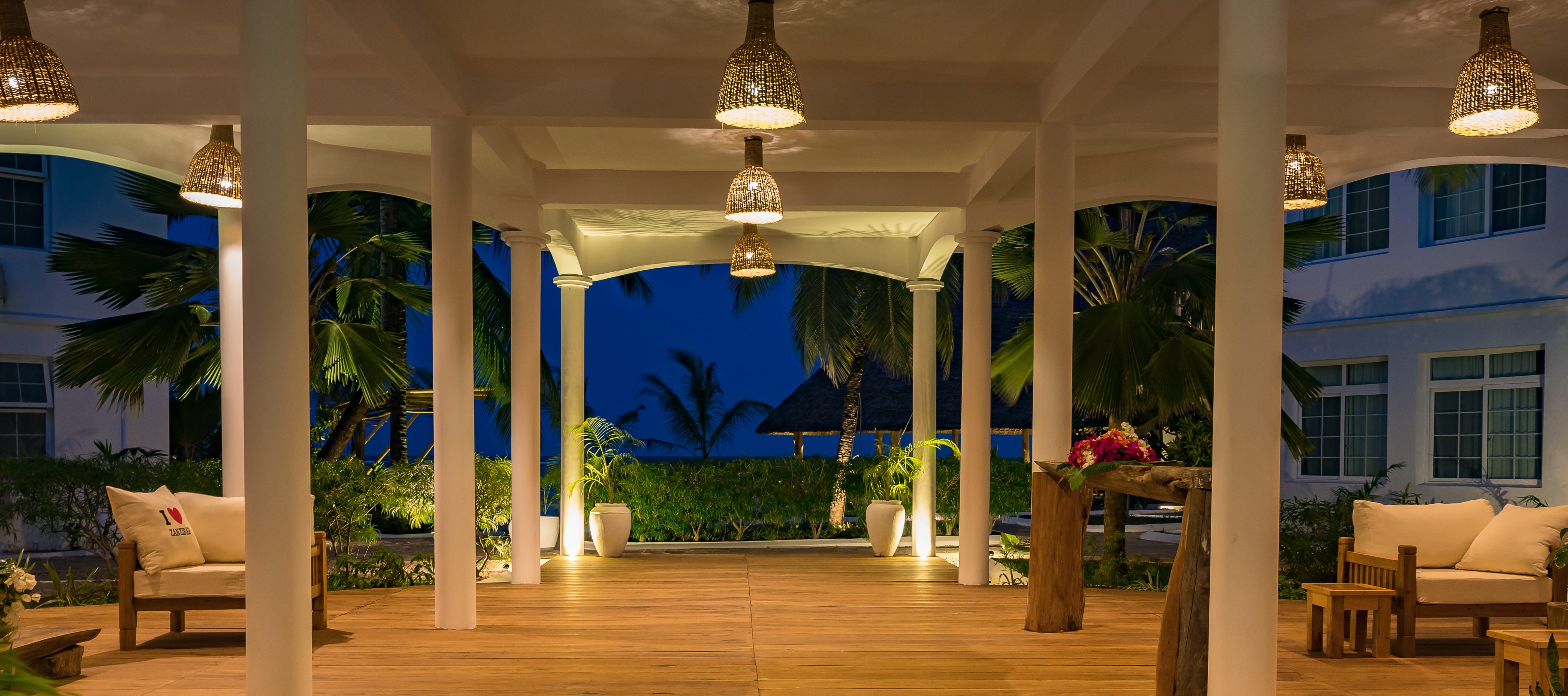 Covered outdoor terrace with teak flooring, white columns, woven lighting, and comfortable seating areas surrounded by tropical landscaping with ocean views