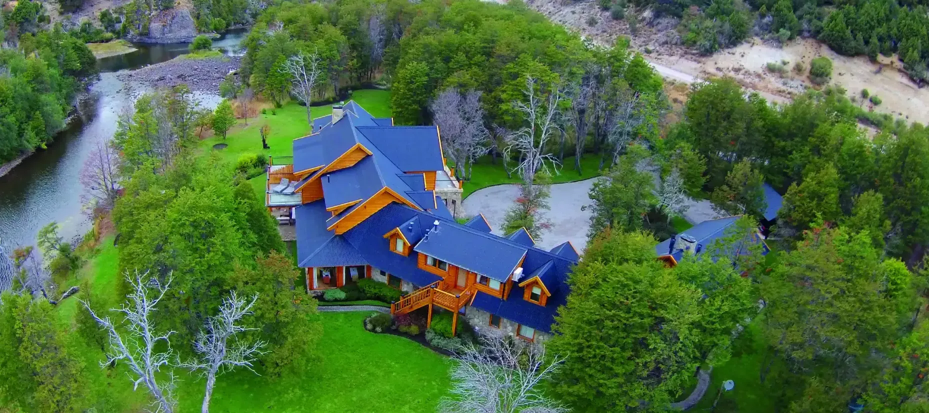 Aerial view of Rio Hermoso Hotel de Montaña nestled in lush Patagonian forest beside a winding river, with mountain peaks rising in the background