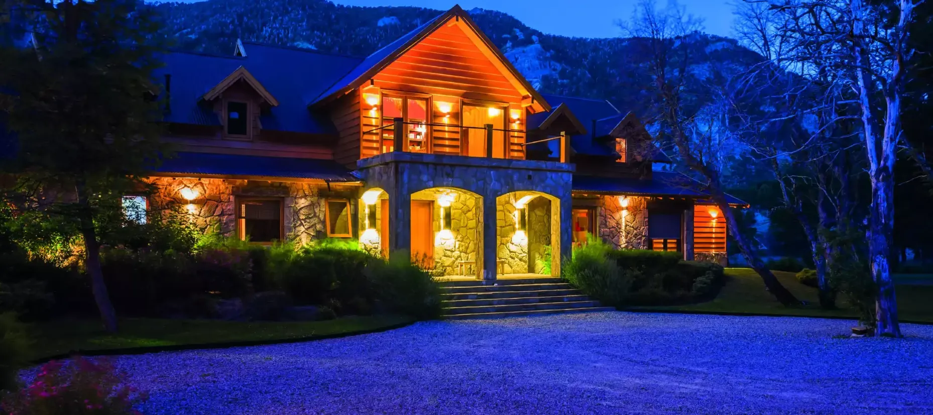 Evening view of Rio Hermoso Hotel de Montaña illuminated with warm lights, showcasing its stone-and-timber facade against a backdrop of forested mountains