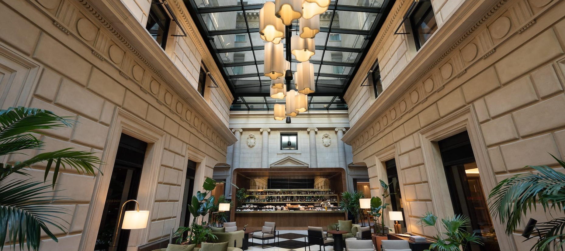 Elegant indoor lounge with tall glass ceiling, modern chandelier, classic stone walls, cozy seating, and a stylish bar at the far end