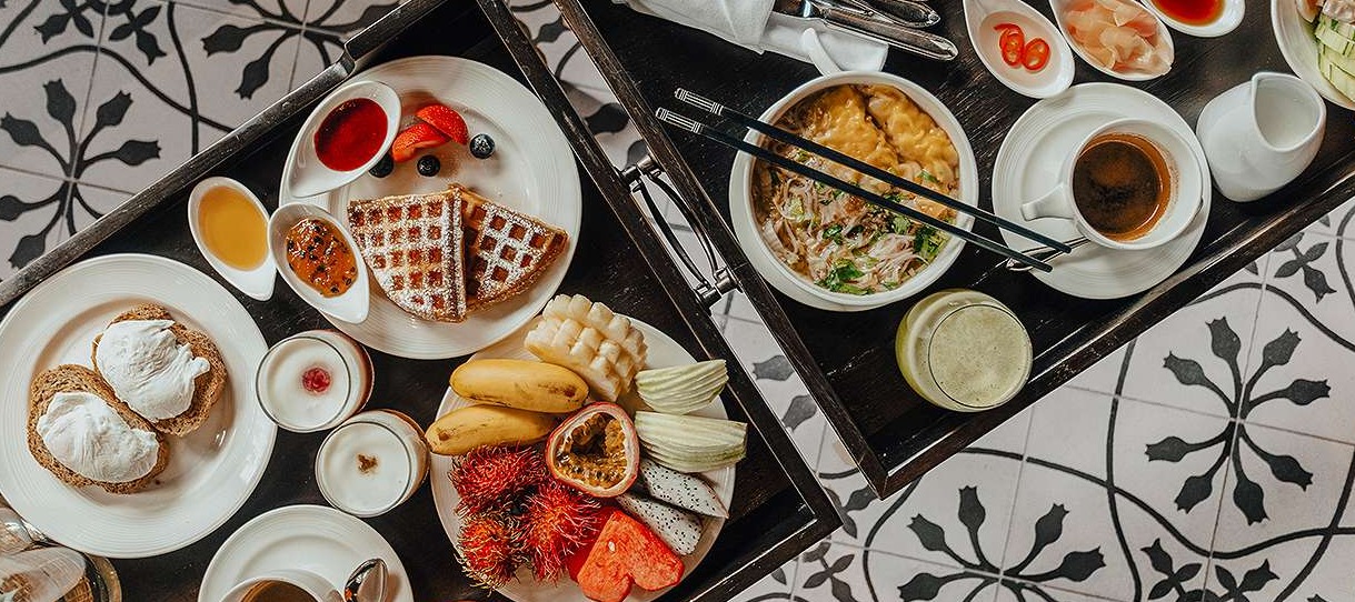Elaborate breakfast spread at 137 Pillars House featuring waffles, tropical fruits, noodle soup, coffee, and various dishes served on white plates atop a dark wooden tray