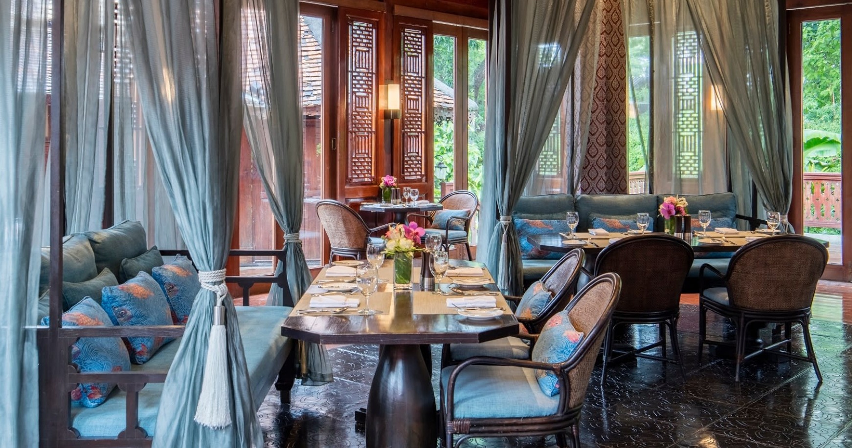 Elegant restaurant interior at 137 Pillars House featuring traditional Thai design with exposed wooden beams, flowing curtains, ornate lanterns, and intimate dining tables set with fine glassware overlooking lush gardens
