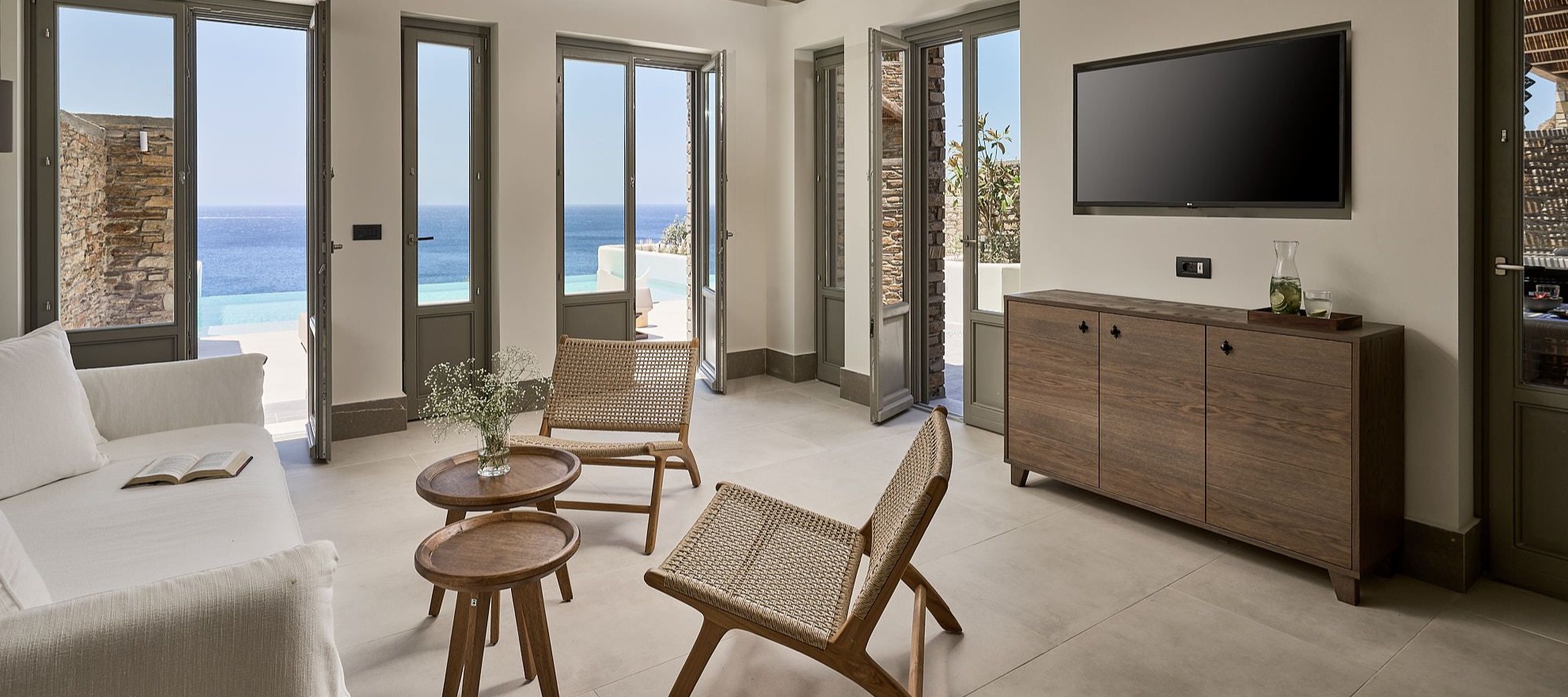 Living room with exposed wooden beams, French doors opening to ocean view, and natural woven furniture