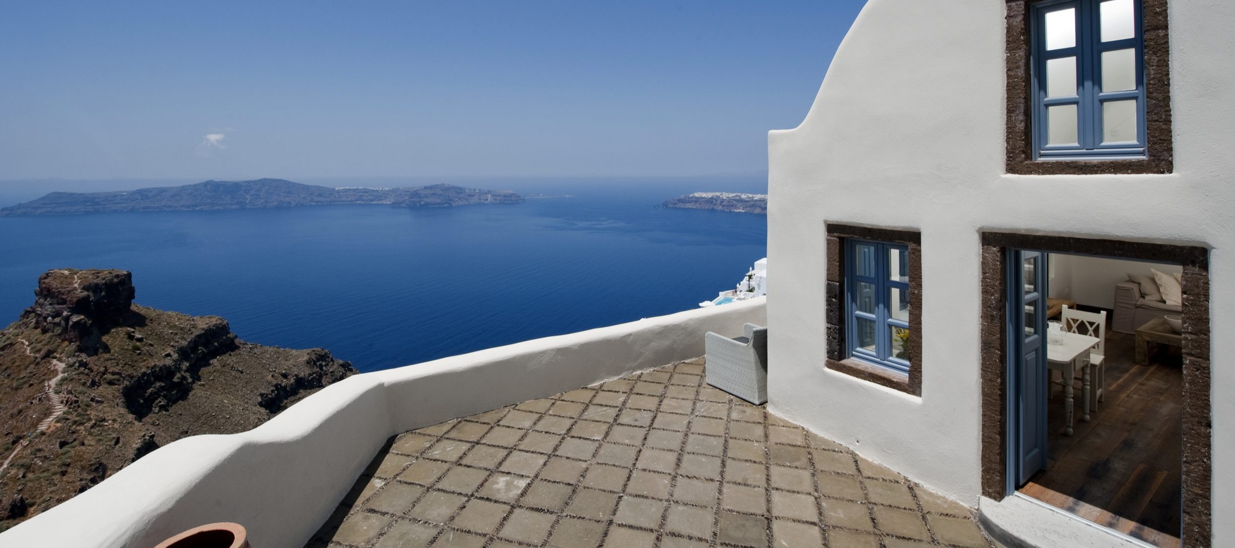 Traditional Greek island terrace with curved white walls, blue shuttered windows, and panoramic view of the Aegean Sea