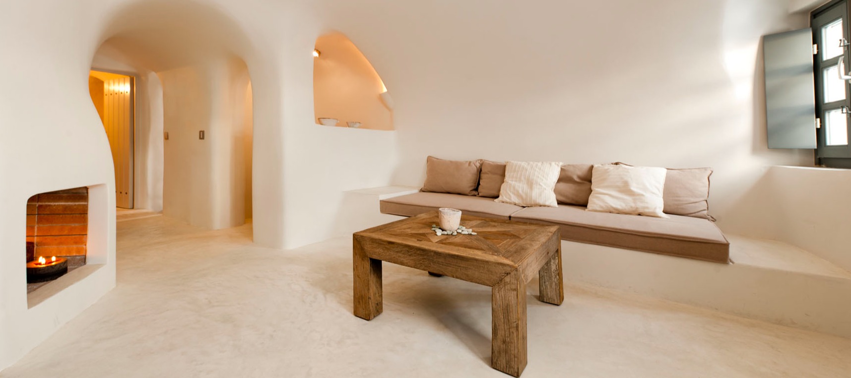 Minimalist Style living room with curved white walls, built-in seating with neutral cushions, rustic wooden coffee table, and traditional fireplace