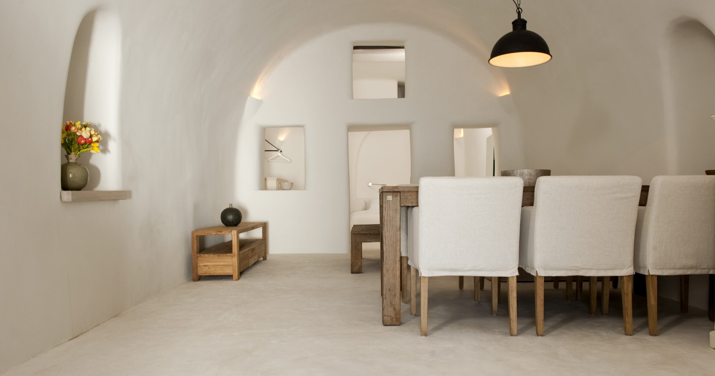 Minimalist dining room with curved white walls, wooden dining table with white upholstered chairs, built-in shelving niches, and black pendant light 