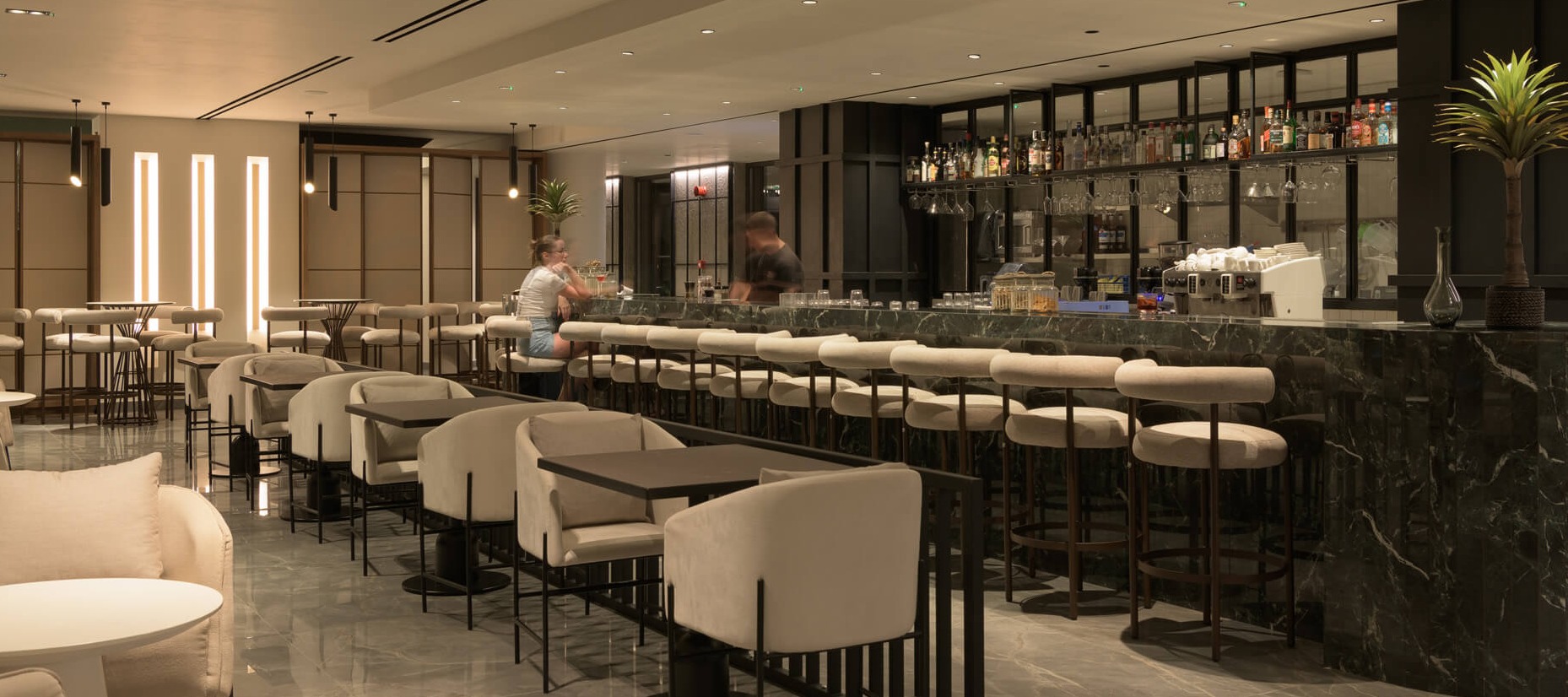 Contemporary hotel bar and lounge with marble surfaces, cream leather seating, linear lighting, and fully stocked bar