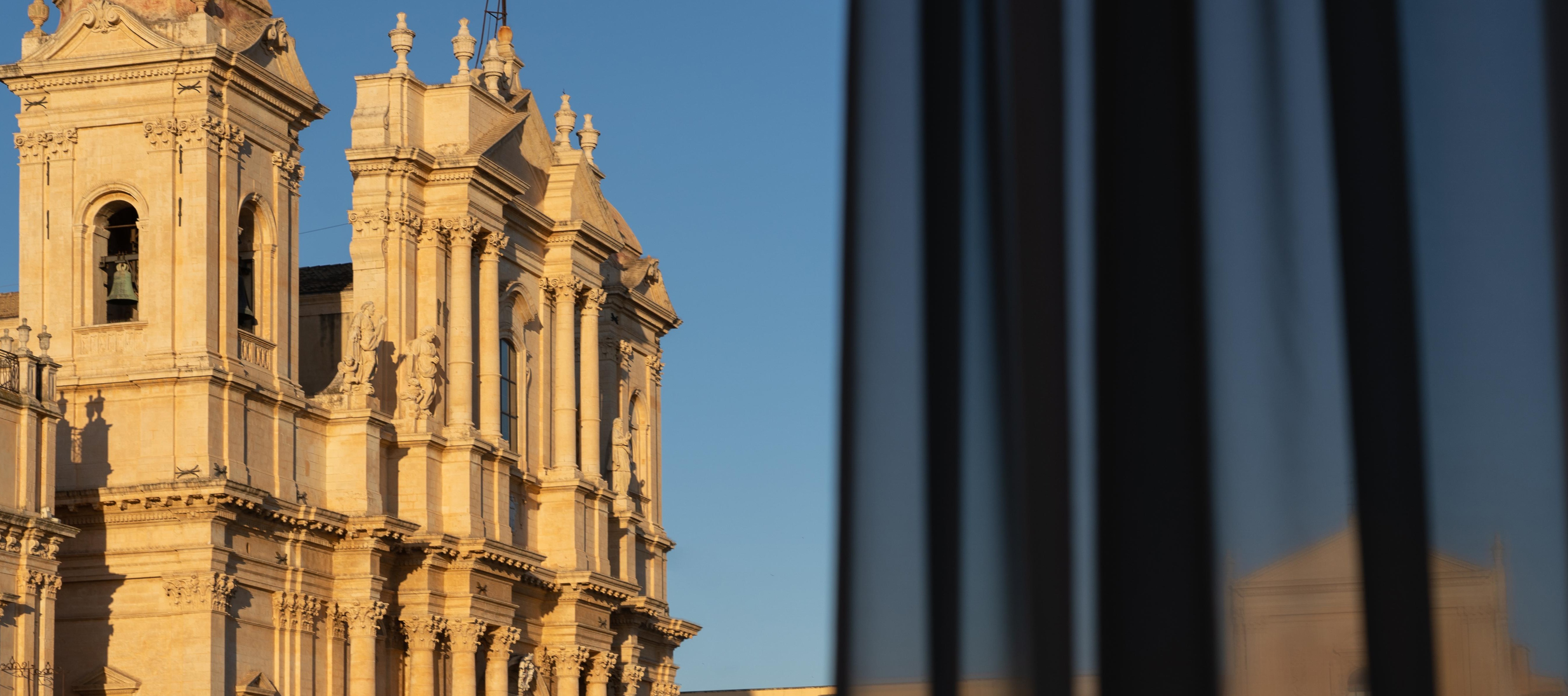 View of the golden Baroque façade of the Noto Cathedral View of the golden Baroque façade of the Noto Cathedral seen from a Q92 hotel room window past a sheer curtain