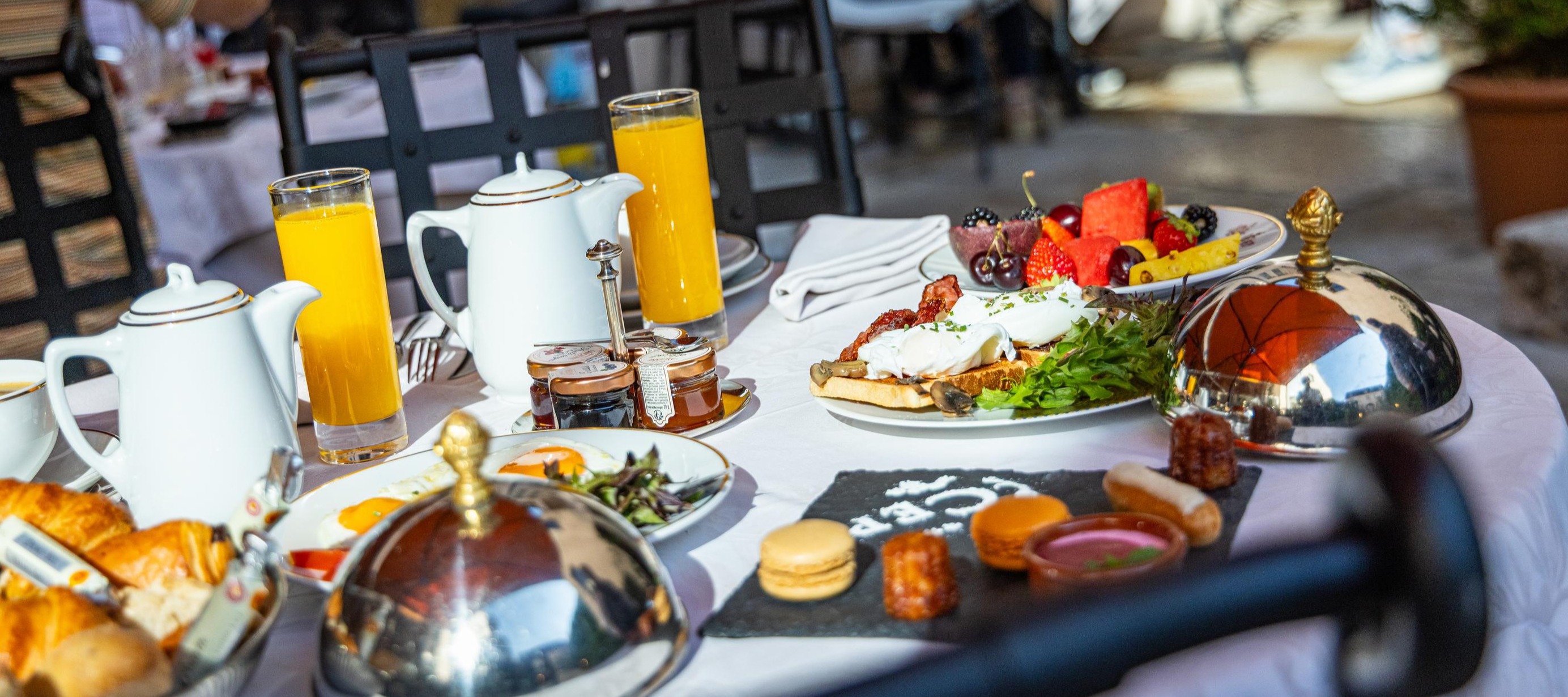 Elegant breakfast spread featuring fresh orange juice, pastries, eggs, fruit platter, macarons, and tea service on an outdoor terrace