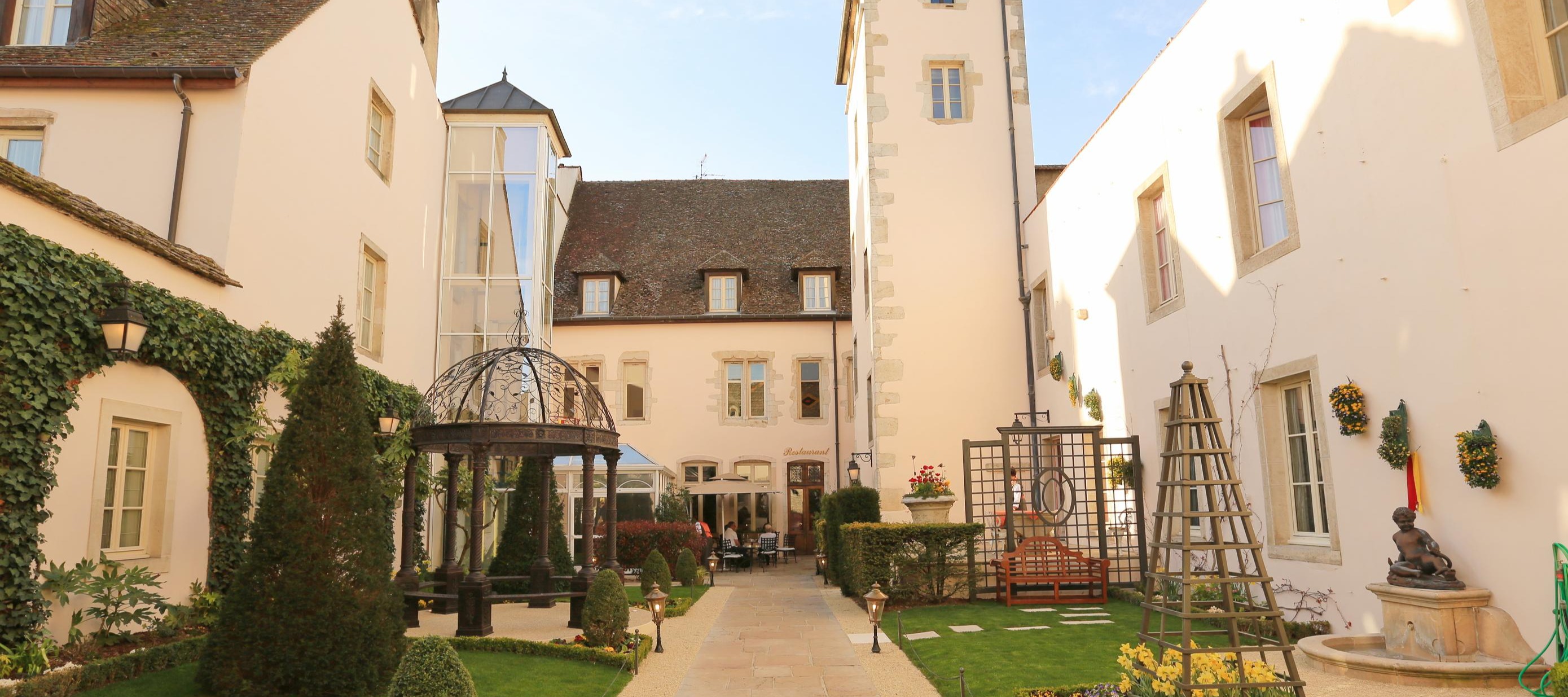 Historic courtyard garden at Hotel Le Cep with cream stone buildings, decorative tower with patterned roof tiles, manicured lawns, and colorful flower beds